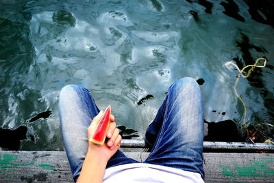 Low section of man with watermelon slice sitting on pier on lake