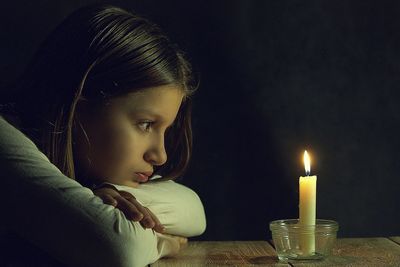 Side view of girl staring at burning candle in bowl on table against black background