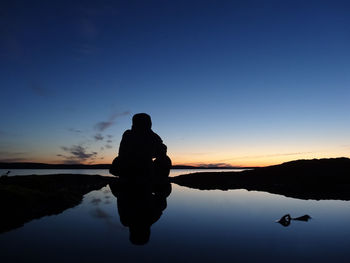 Silhouette person sitting on wet field against sky during sunset