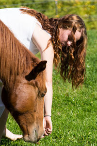 Young woman feeding horse while standing on grassy field during sunny day
