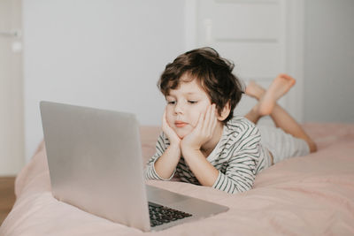 Little boy lie in the bedroom on the bed and look at the laptop. high quality photo