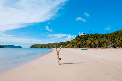 Woman practicing handstand at beach against blue sky