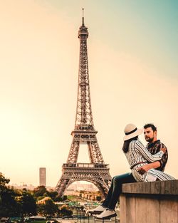 Couple romancing while sitting on retaining wall against eiffel tower during sunset