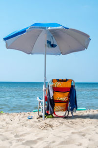 An empty beach chair under a big blue umbrella, is draped with clothing, near  lake michigan