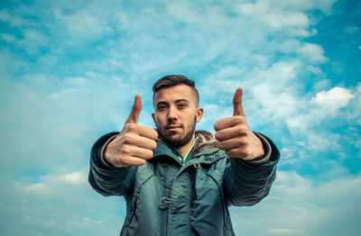 Portrait of young man showing thumbs up sign against cloudy sky