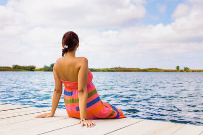 Rear view of woman sitting on pier over lake against sky