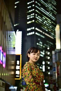 Portrait of smiling young woman standing against illuminated built structure