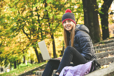 Girl with wool cap sitting in the park using laptop. teen use computer to communicate with friends 