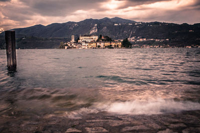 Mid distance of buildings at riverbank by mountains against cloudy sky