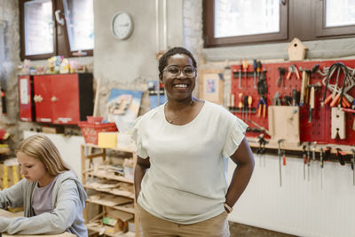 Portrait of happy female teacher standing with hands in pockets next to girl at school workshop