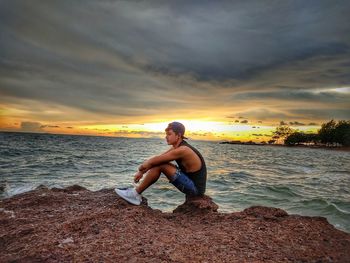 Woman sitting on rock at beach against sky during sunset