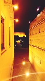 Illuminated street by buildings against sky at night
