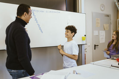 Smiling boy talking to male teacher standing near whiteboard in classroom