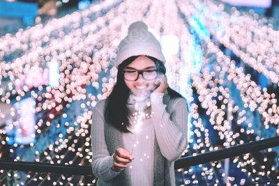 Young woman burning sparkler while standing against illuminated lights