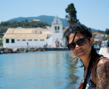 Tattooed woman in sunglasses standing by lake during sunny day