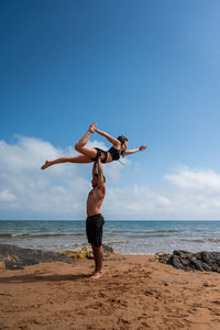 Full body shirtless man standing on sandy beach near waving sea and holding barefoot girlfriend in raised arms against cloudy blue sky on sunny summer day