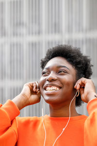 Happy african american female with black hair listening to music in earphones while standing on street near wall of city looking away