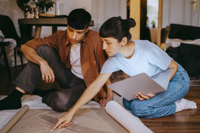 Young female freelancer pointing at blueprint while discussing with male colleague sitting on floor at home office