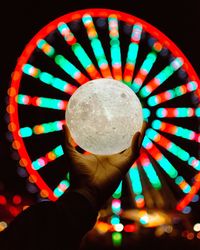 Close-up of person holding illuminated lighting equipment against ferris wheel