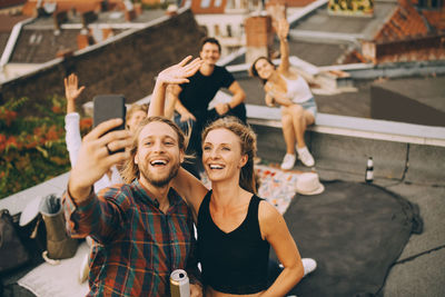 Cheerful man taking selfie with friends on smart phone while enjoying party on terrace