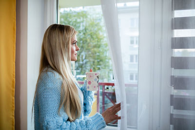 A young woman stands and looks out the window at the morning outside.