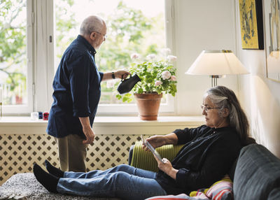 Full length of senior woman using digital tablet while man watering flower plant at home