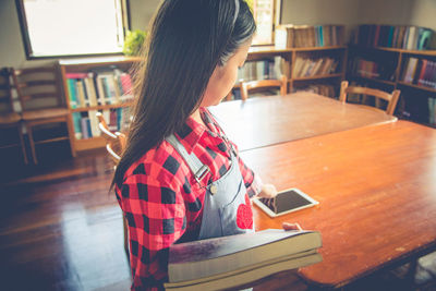 Girl holding book while taking digital tablet from table in library