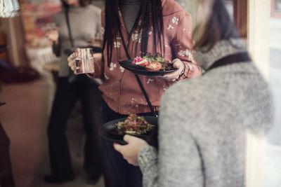 Female colleagues eating lunch while standing at workshop seen from window glass