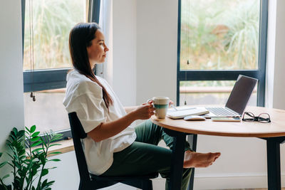 Young female freelancer drinking coffee and working on laptop at home.