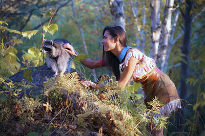 Side view of young woman in traditional clothing stroking raccoon in forest