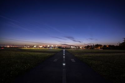 Empty road on illuminated field against sky at dusk