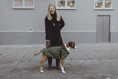 Cheerful young woman standing with boxer against building in city