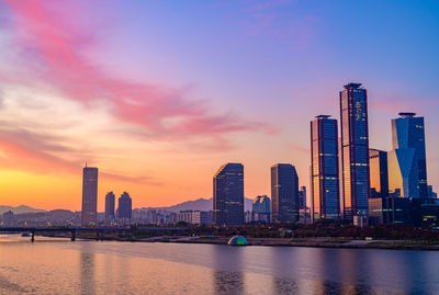 Sea by modern buildings against sky during sunset