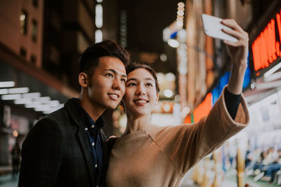 Smiling couple doing selfie while standing on street in city at night