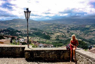 Portrait of woman sitting on retaining wall at observation point against landscape