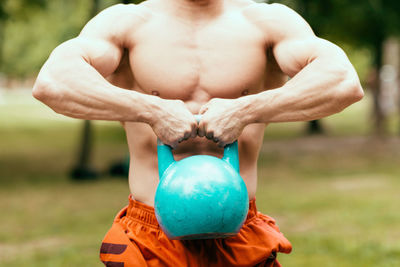 Midsection of shirtless man lifting kettlebell on field