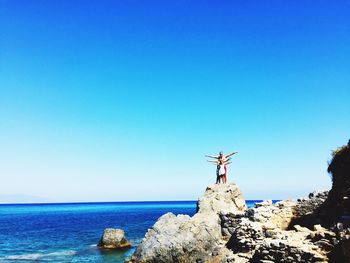 Family enjoying on rock by sea against clear blue sky