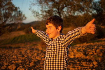 Smiling boy with arms outstretched standing on land during sunset
