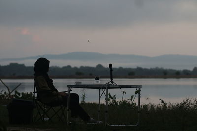 Silhouette man sitting on chair by lake against sky during sunset