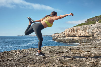 Full length of young woman on rock at sea shore against sky