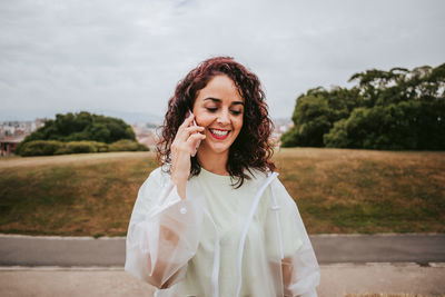 Smiling young woman using phone while standing against sky