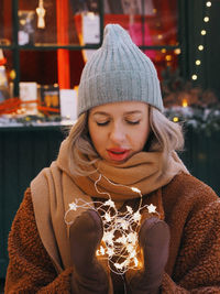 Young blonde woman wearing warm clothes holding garland posing at christmas street market 