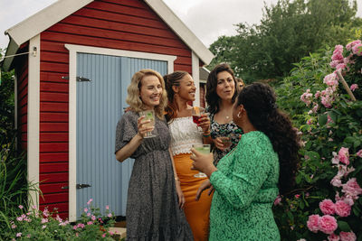 Female friends holding drinks while talking to each other standing in garden