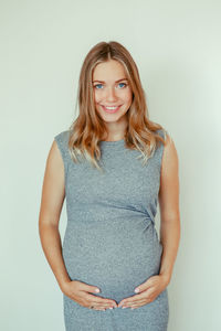 Portrait of pregnant young woman touching abdomen while standing against white background