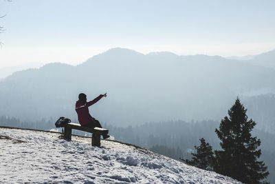 Woman pointing while sitting on bench at snowcapped mountain