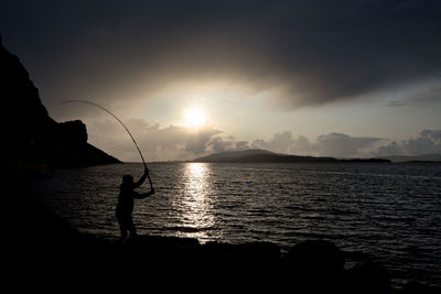 Silhouette man fishing in sea against sunset sky