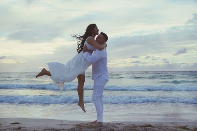 Side view of man lifting woman on shore at beach during sunset