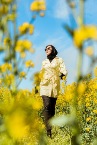A young stylish woman poses among a blooming yellow field of rapeseed under a bright spring sky