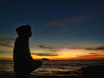 Silhouette man crouching on beach against sky during sunset