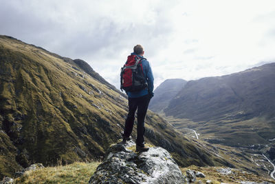 Rear view of man with backpack standing on rock against sky